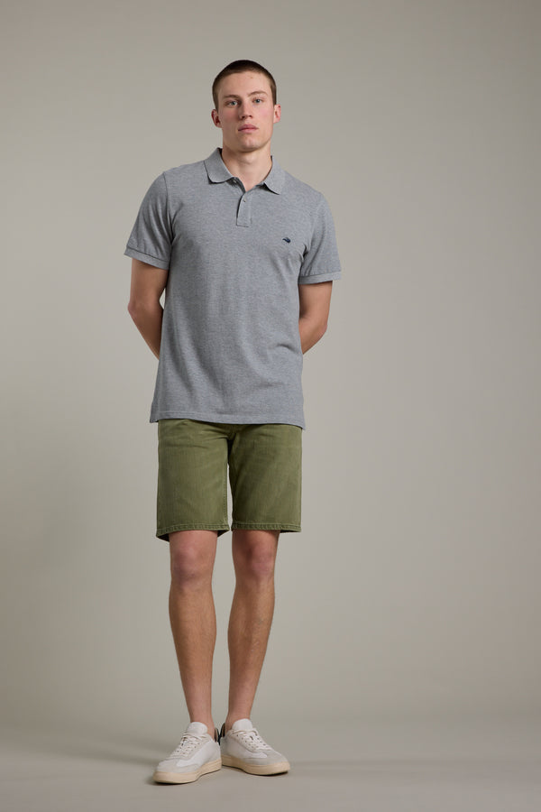 A young man stands against a plain background wearing the Barkers Regent Pique Polo with olive green shorts and white sneakers, his hands behind his back and a neutral expression.