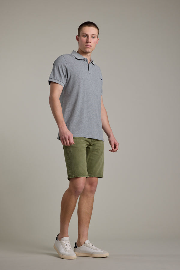 A young man stands against a plain background, wearing Barkers Rover Coloured Denim Shorts made from regenerative cotton, a gray polo shirt, and white sneakers. He faces slightly to the side with a neutral expression.