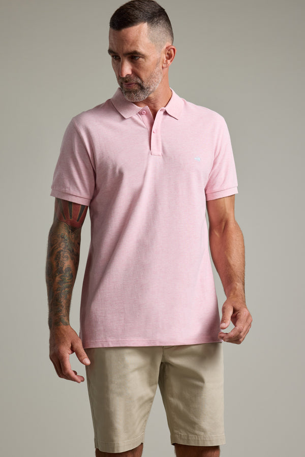 Man in a light pink Barkers Regent Pique Polo and beige shorts, standing against a plain backdrop.