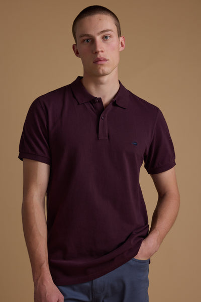 A young man with short hair wears a maroon Barkers Regent Pique Polo and blue pants, standing against a plain beige background with one hand in his pocket, looking directly at the camera with a neutral expression.