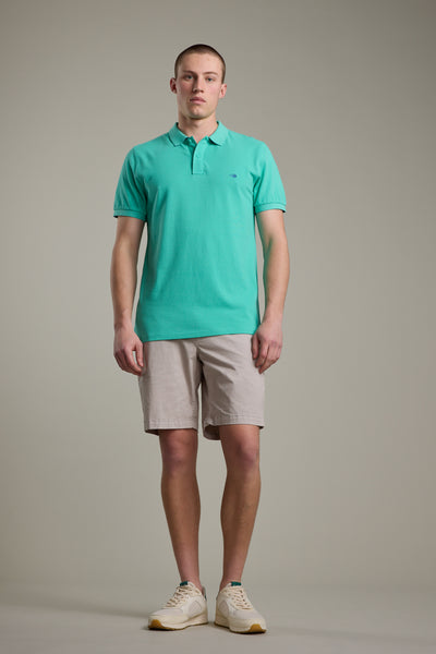 A person stands against a plain background wearing a light turquoise Barkers Regent Pique Polo made from organic cotton, beige shorts, and white sneakers. They have short hair, a neutral expression, and relaxed arms by their sides.