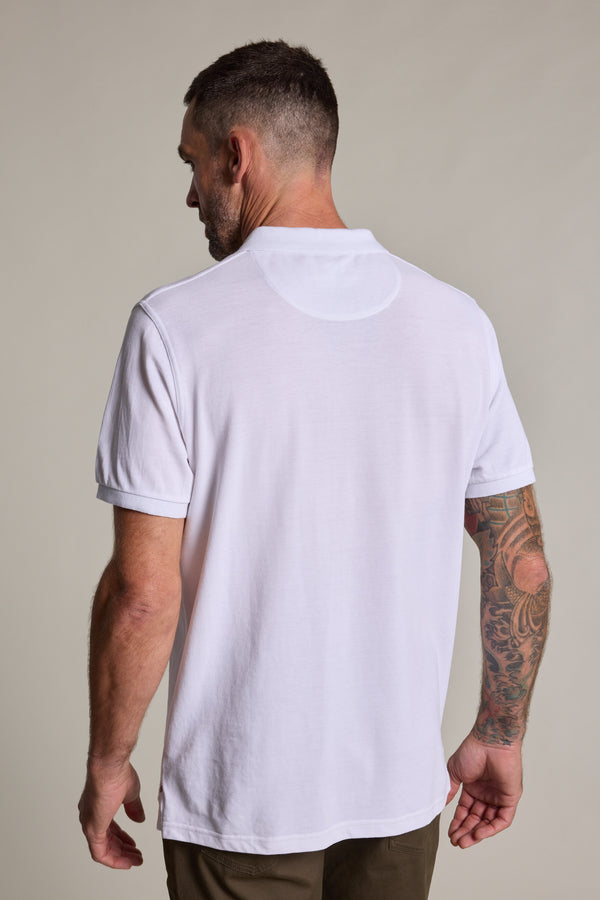 A man with short hair and a tattooed right arm stands with his back to the camera, wearing a white Barkers Regent Pique Polo and olive-green pants against a plain background.