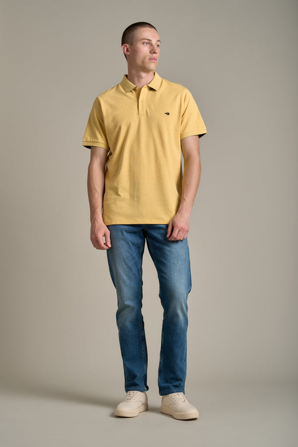 A man stands before a plain background wearing a yellow Regent Pique Polo by Barkers, blue jeans, and beige sneakers. He looks slightly to his left with his arms relaxed at his sides.