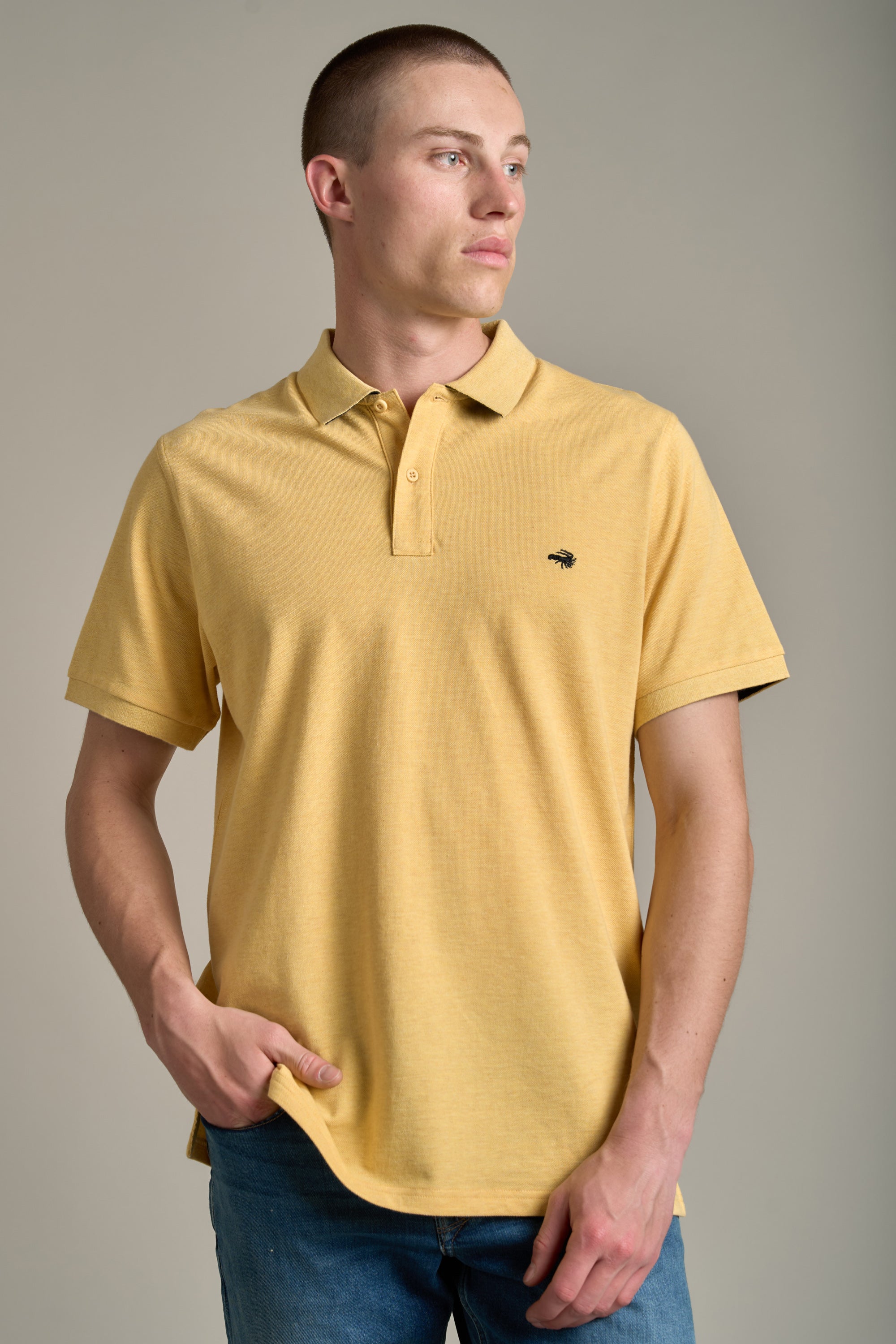 A man in a light yellow Barkers Regent Pique Polo and blue jeans stands against a plain beige background, looking to his left with one hand in his pocket.