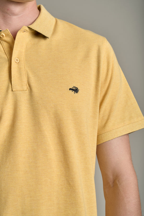 A person wears a light yellow Barkers Regent Pique Polo in organic cotton featuring a small black embroidered crayfish logo on the chest. The image shows their upper torso and part of an arm against a plain background.