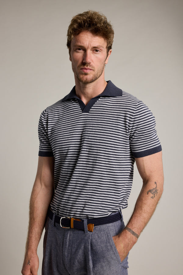 A man with short, curly hair and a trimmed beard faces the camera in the Barkers Parkway Cotton Stripe Polo, made from responsibly sourced cotton, paired with blue trousers and a belt, revealing a tattoo on his left forearm.