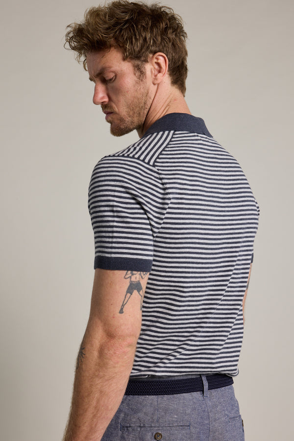 A man with short, curly hair and a tattoo on his upper arm stands sideways, looking down. He wears the Barkers Parkway Cotton Stripe Polo, crafted from responsibly sourced cotton, paired with blue pants against a plain background.