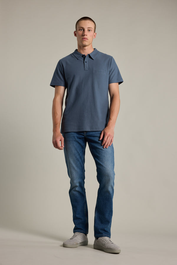 A person stands facing forward against a plain light background, wearing the Barkers Canal Polo (short-sleeve, blue), blue jeans, and light gray sneakers.