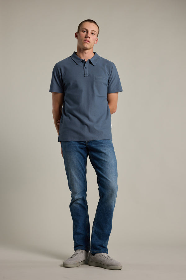 A person stands against a plain background, wearing a blue Barkers Canal Polo, blue jeans, and gray sneakers, with arms behind their back and a neutral expression.