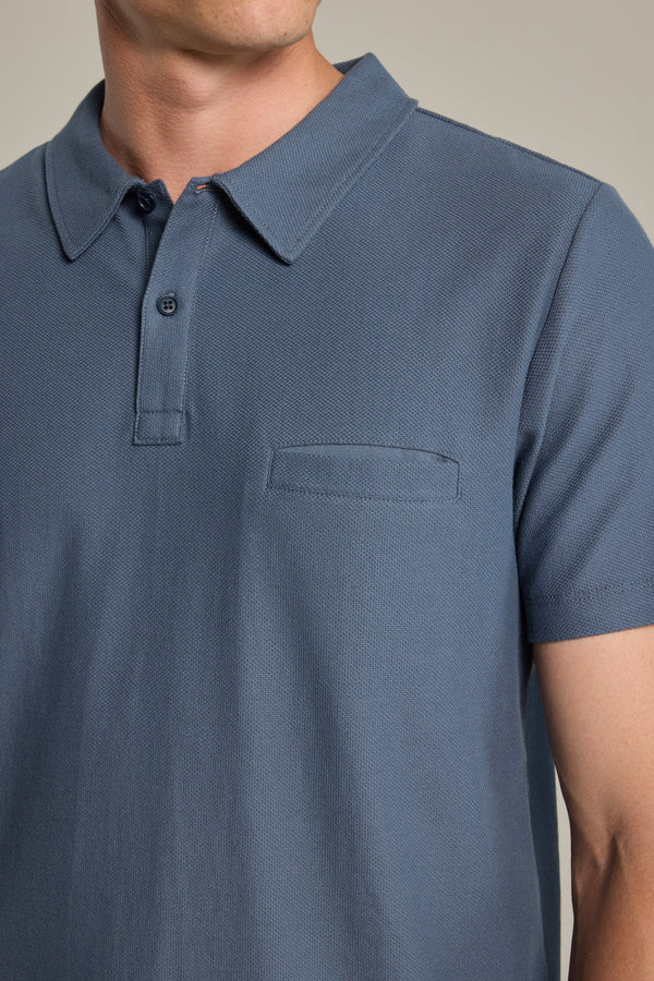 A person wearing the Barkers Canal Polo, a short-sleeved blue Supima cotton shirt with a button placket and small horizontal chest pocket, photographed from shoulders to waist.