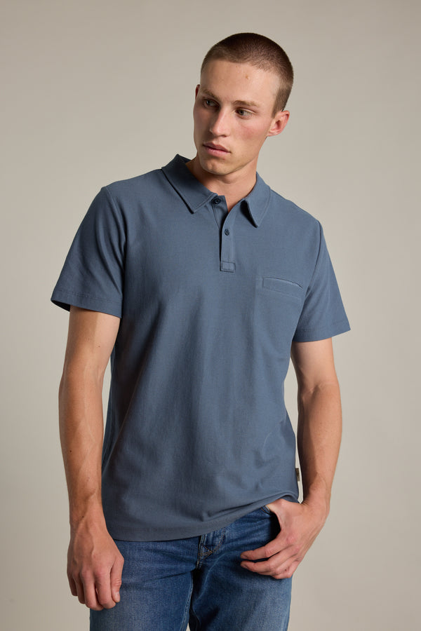 A young man with short hair wears a Barkers Canal Polo in blue and blue jeans, posing against a plain light background with one hand in his pocket and a neutral expression.