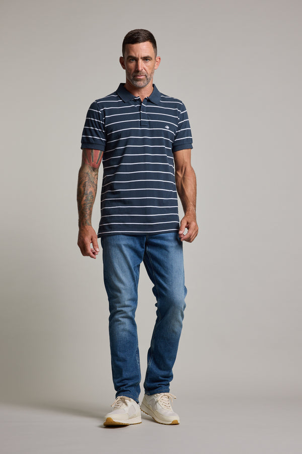 A man with short dark hair and a tattooed right arm wears the Barkers Regent Stripe Polo in navy and white, paired with blue jeans and white sneakers, standing against a plain light gray background.