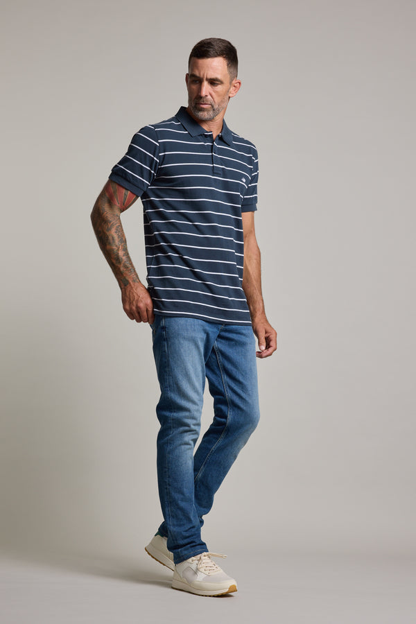 A man with short hair and a tattooed left arm wears the Barkers Regent Stripe Polo, navy blue and white, made from organic cotton, paired with blue jeans and white sneakers, standing against a plain light gray background.