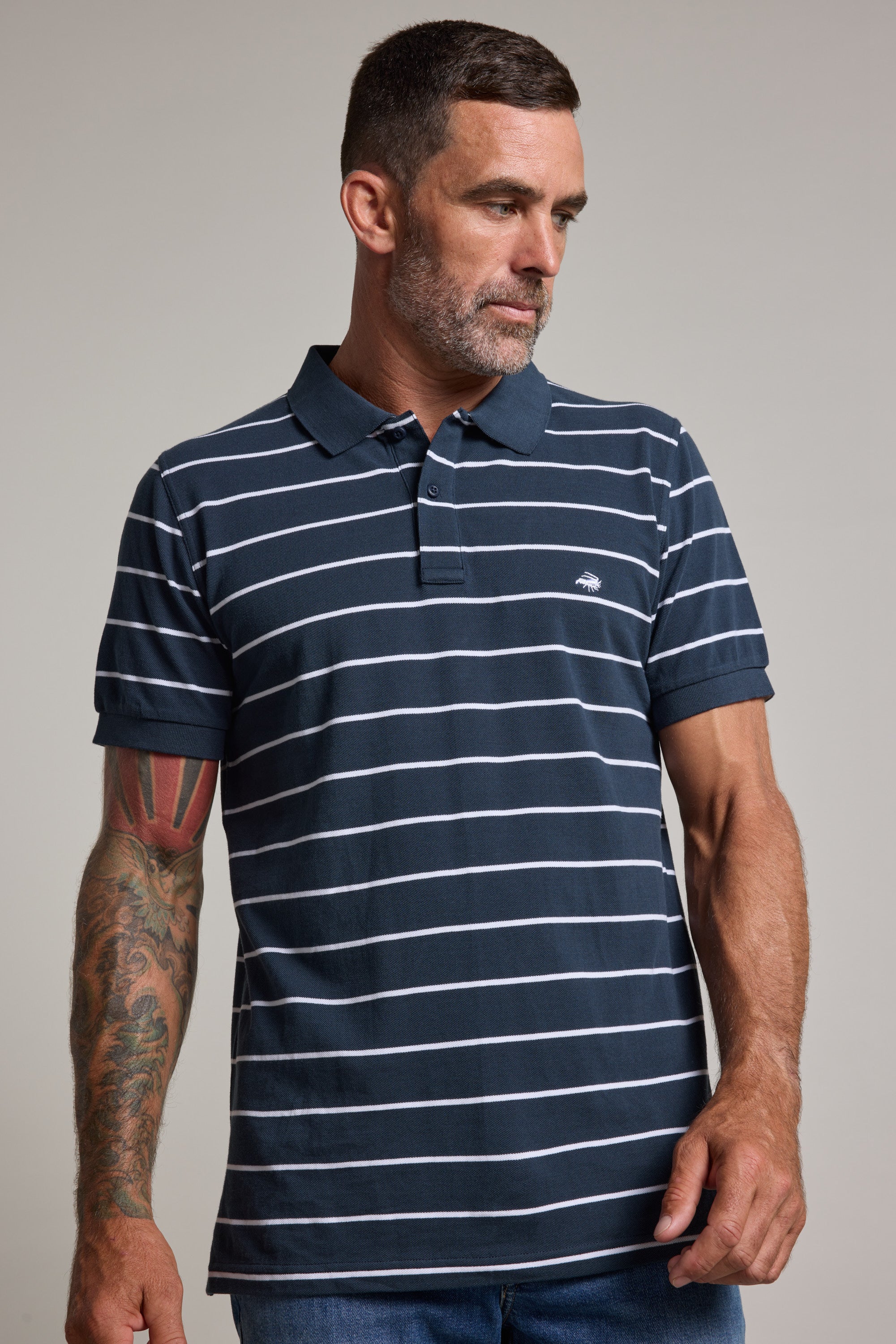 A man with short dark hair and a beard wears the Barkers Regent Stripe Polo, a navy blue short-sleeve polo shirt with white horizontal stripes. He has tattoos on his left forearm and looks slightly down and to his left.