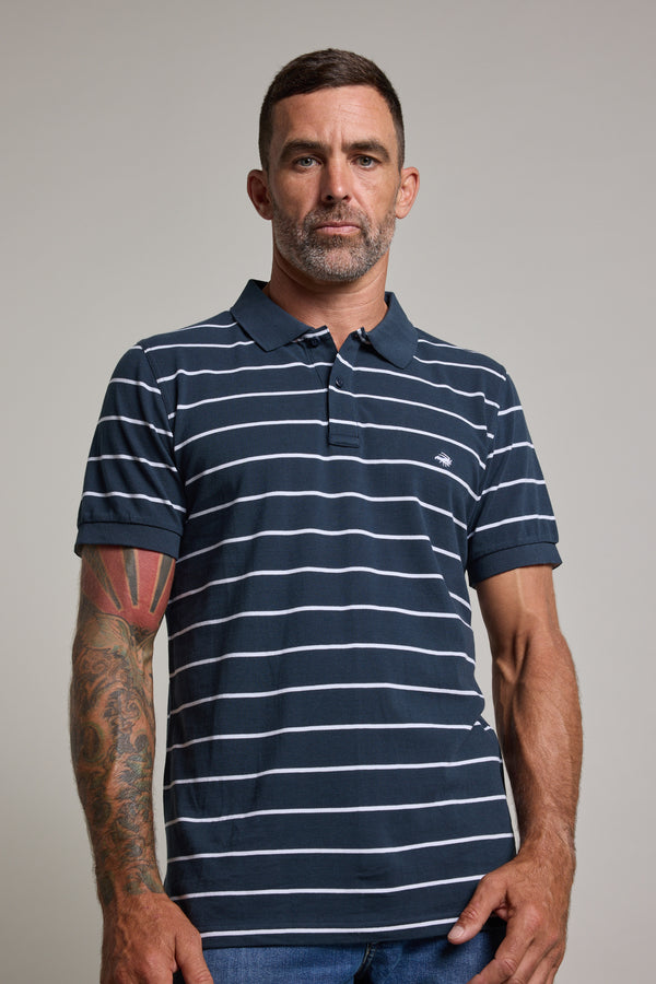 A man with short dark hair and a beard wears a Barkers Regent Stripe Polo in navy and white. Made from organic cotton, the shirt is showcased as he stands against a plain background, revealing his tattooed left arm and neutral expression.