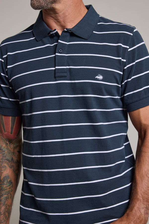 A man with a tattooed left arm and partially visible face wears a Barkers Regent Stripe Polo, a navy blue limited edition polo with white stripes and a small embroidered chest logo.
