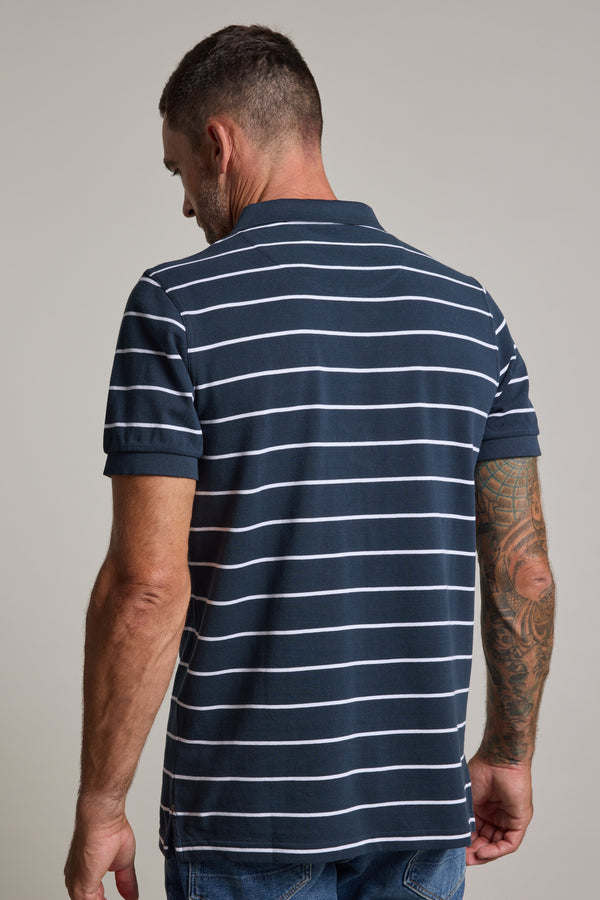 A man with short hair and visible right arm tattoos stands with his back to the camera, wearing the Barkers Regent Stripe Polo— a navy blue, short-sleeved shirt with white stripes— and blue jeans.