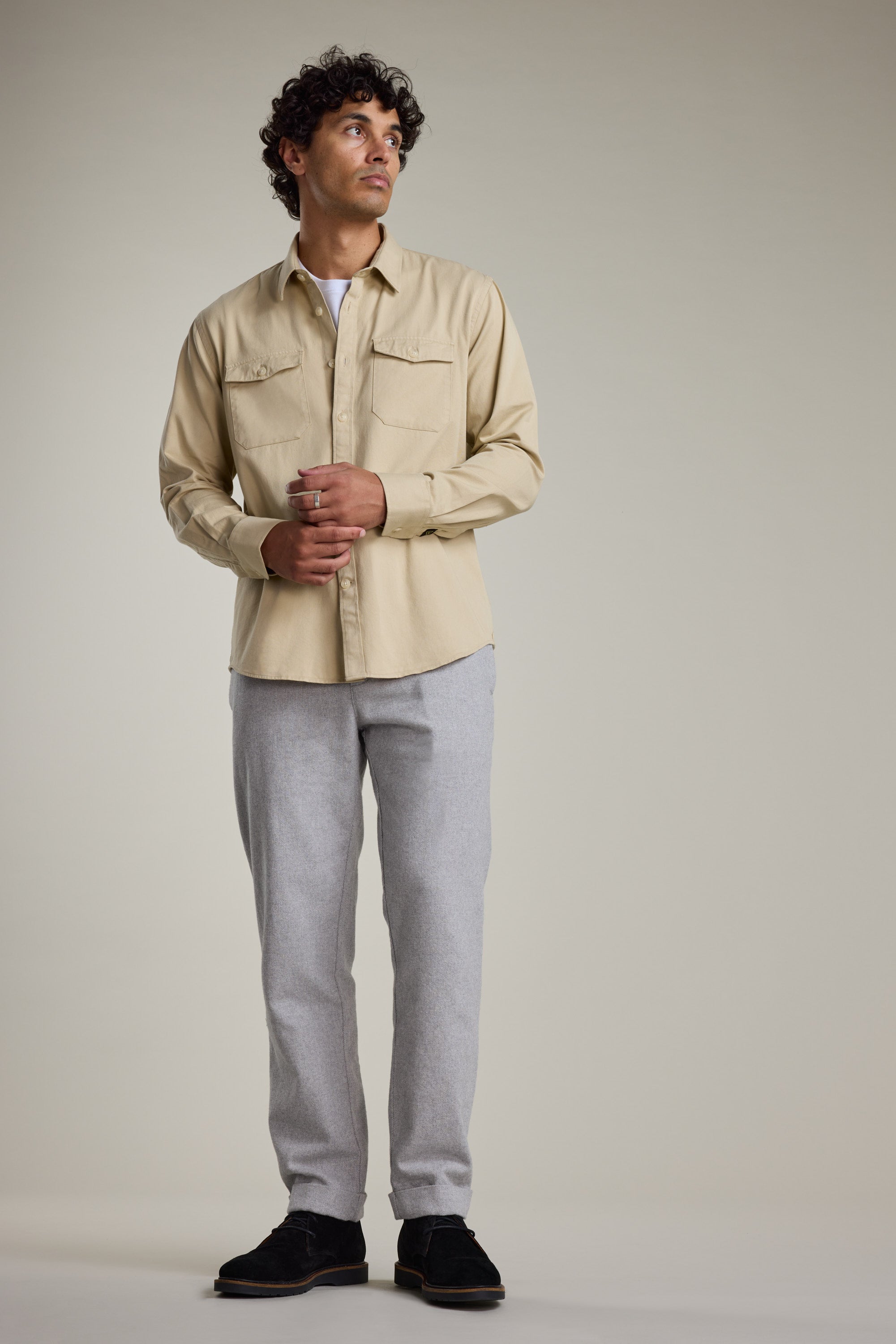 A man with curly hair stands against a plain background, wearing a beige shirt over white, black shoes, and Barkers Dunmore Pant—light gray smart-casual trousers crafted from responsibly sourced cotton. He gazes upward with hands loosely clasped.