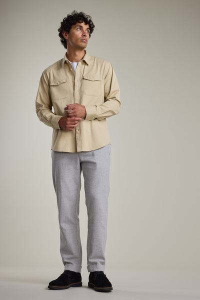 A man with curly hair stands against a plain background, wearing a beige shirt over white, black shoes, and Barkers Dunmore Pant—light gray smart-casual trousers crafted from responsibly sourced cotton. He gazes upward with hands loosely clasped.