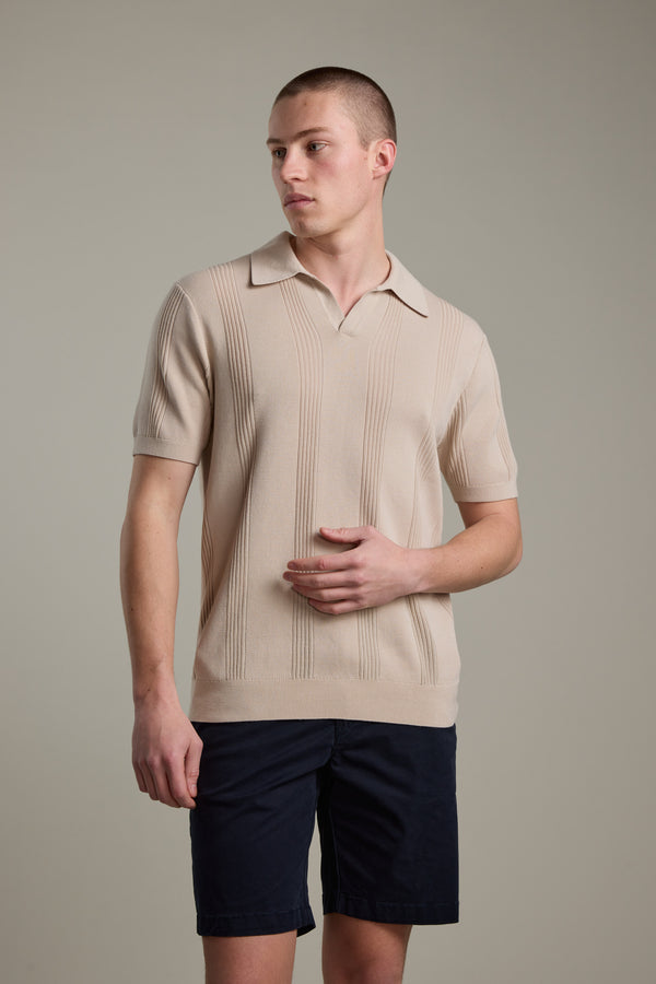 A man in the Barkers Milwood Knit Polo and dark navy shorts stands against a light gray background, gazing left with one hand on his torso—a summer essential for effortless style.