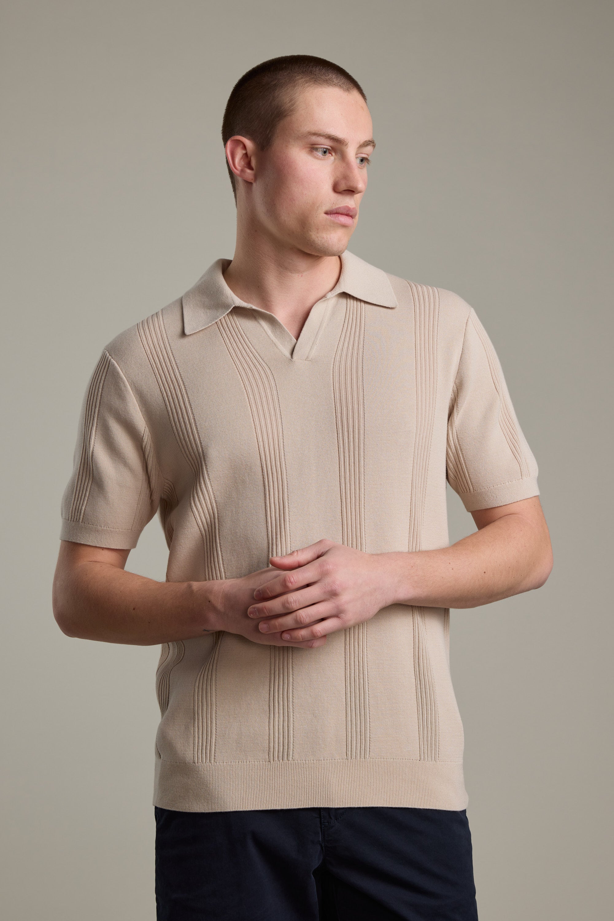 A man with short hair stands against a light gray background, wearing the Barkers Milwood Knit Polo in beige and dark pants, looking to the side with his hands loosely clasped in front of him.