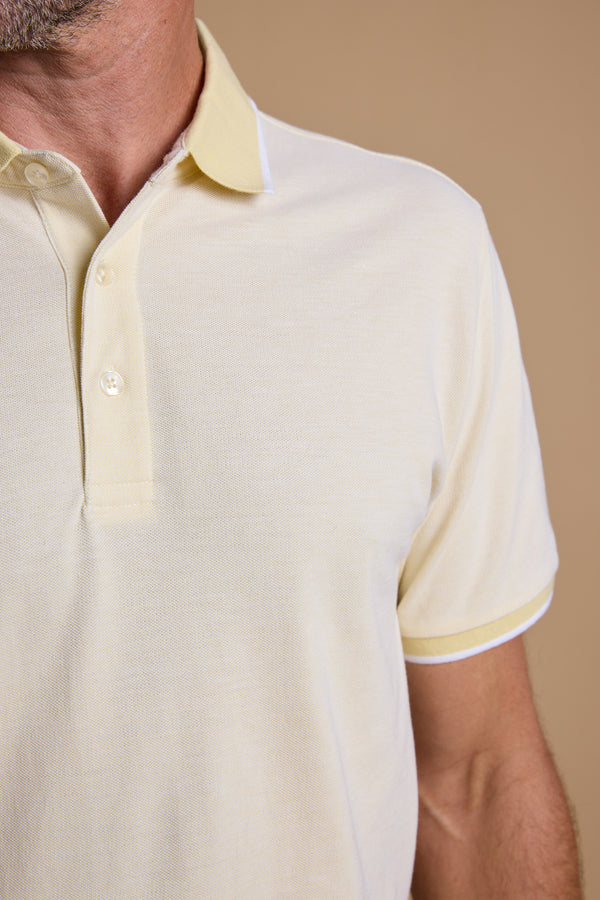 A close-up shows a man wearing the Barkers Birdie Pique Polo in light yellow, featuring a collar and three buttons, against a beige background. Only his lower face, neck, and upper torso are visible.