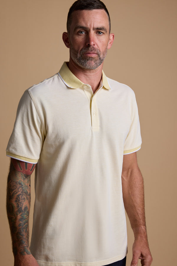 A man with short hair, a beard, and a sleeve tattoo on his left arm wears the Barkers Birdie Pique Polo in light yellow, standing against a plain beige background.