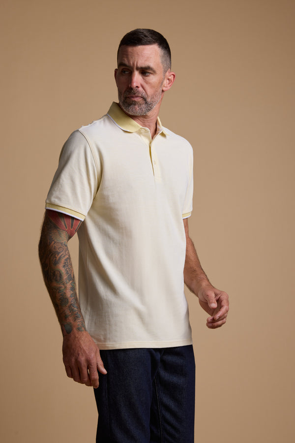 A man with short dark hair and a beard wears the Barkers Birdie Pique Polo in yellow with dark pants, standing against a beige background. He has tattoos on his left forearm and looks slightly to his right.