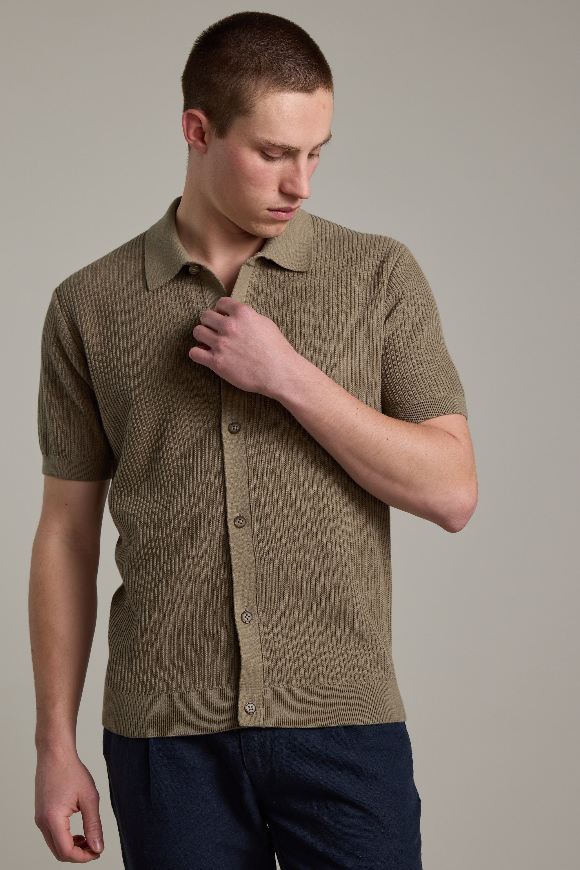 A man wearing the Barkers Barrows Knit Button Up Polo—a short-sleeve, ribbed, khaki shirt made from responsibly sourced cotton—pairs it with dark pants and stands against a neutral gray background.
