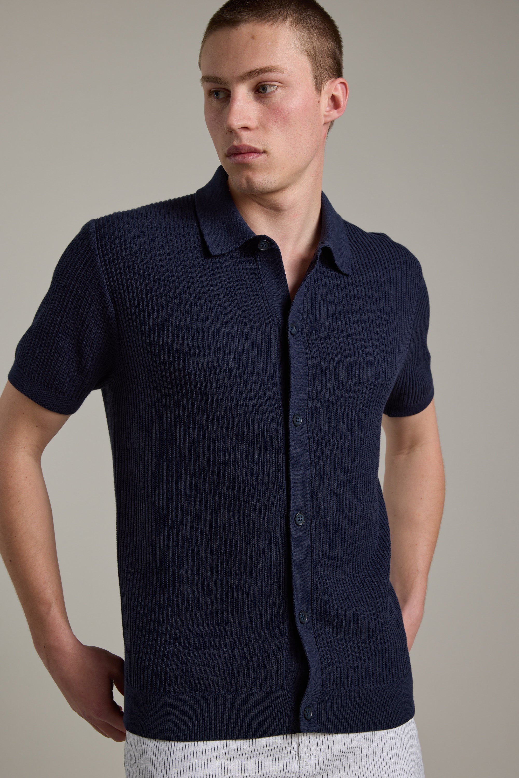 A man stands against a plain background, looking left with one hand on his hip, wearing the Barkers Barrows Knit Button Up Polo in navy blue cotton with short sleeves and a spread collar.
