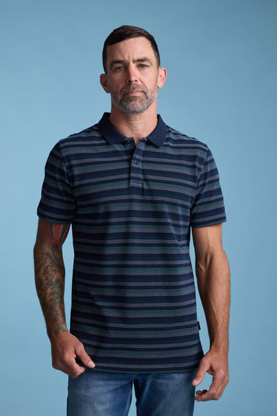 A man with short dark hair and a trimmed beard models the Barkers Dalton Stripe Polo with jeans. His colorful right-arm tattoo stands out against a solid light blue background—ideal for a stylish summer wardrobe.