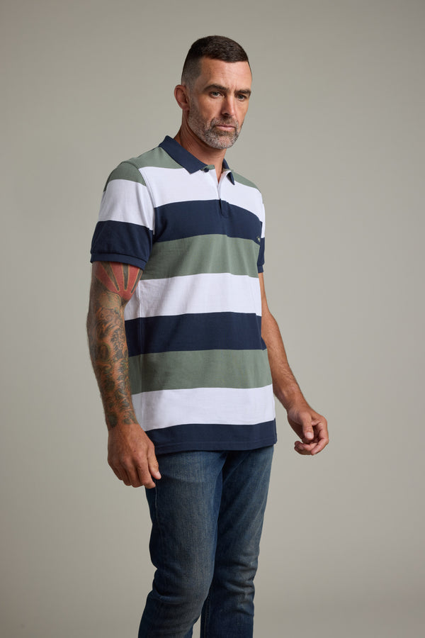A man with short dark hair and a beard wears the Barkers Regent Pique Stripe Polo in green, white, and navy stripes with blue jeans. His visible left-arm tattoos stand out against a plain gray background.