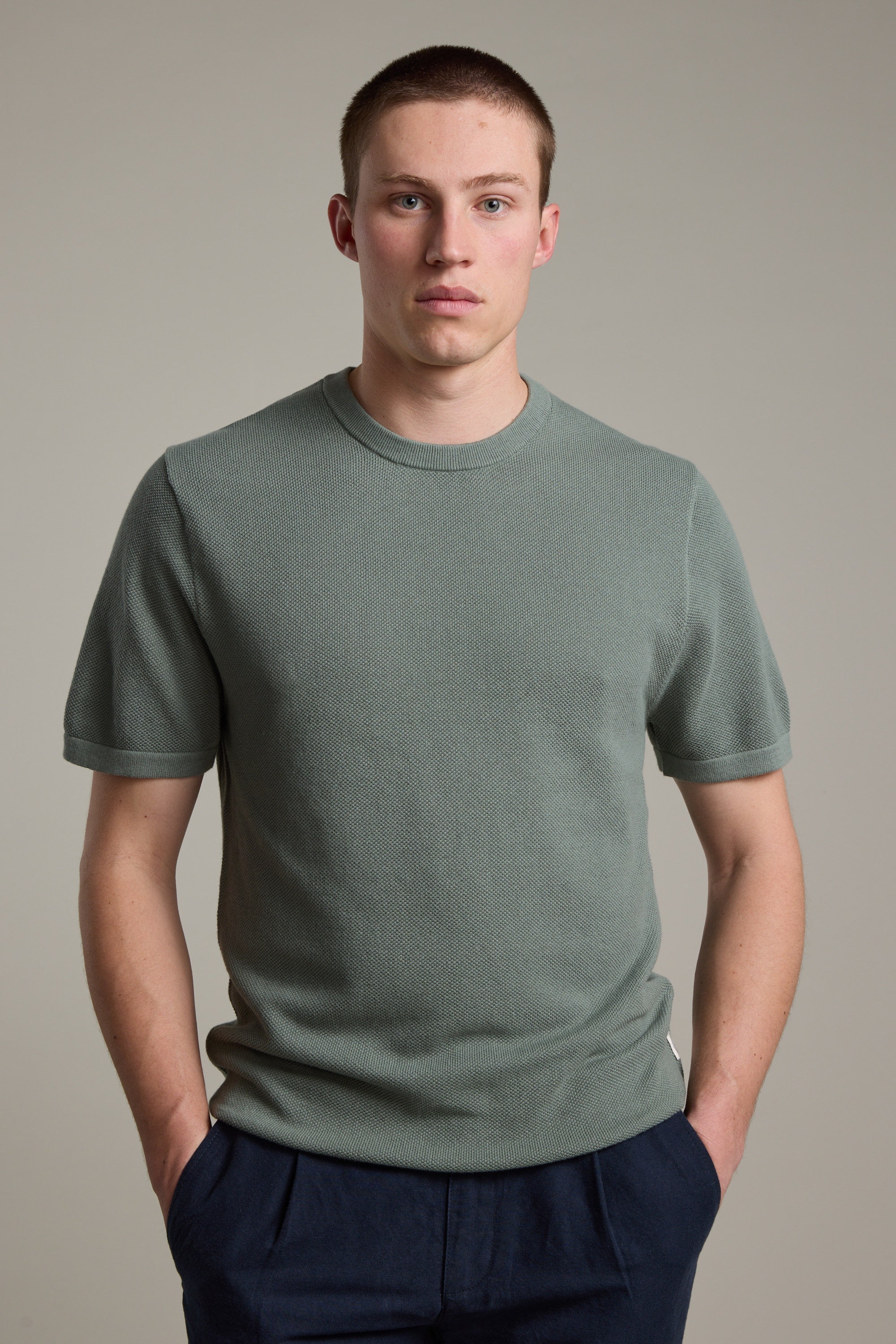 A young man with short brown hair wears the Barkers Flinders Cotton Knit Tee in muted green and dark pants. He stands against a plain light gray background, hands in pockets, looking directly at the camera.