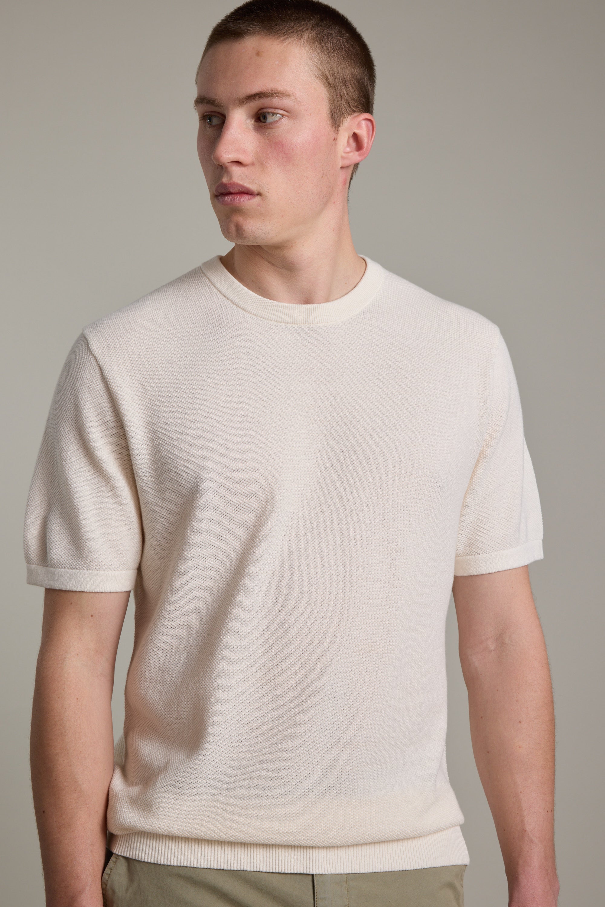A young man with short hair wears the Barkers Flinders Cotton Knit Tee, a cream organic cotton crewneck, paired with light pants. He stands against a neutral background, gazing slightly to his left.
