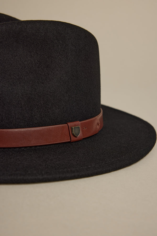 The Brixton Messer Fedora Hat by Brixton is a black wool felt wide-brim hat with a brown leather band, an adjustable strap, and a small metallic shield-shaped emblem on the side.