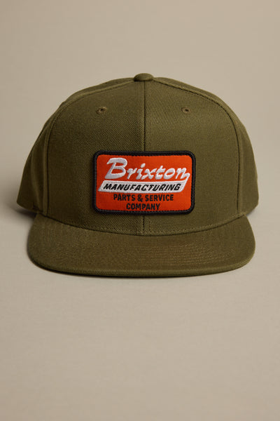 The Brixton Township MP Snapback Cap by Brixton is an olive green 6-panel hat with a flat brim, featuring a red and white front patch that reads "Brixton Manufacturing Parts & Service Company," plus an adjustable snapback closure.