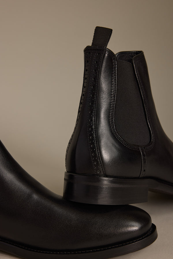 Close-up of two Barkers Peter Chelsea Boots in black premium leather with elastic side panels, detailed stitching, and a pull tab, highlighting quality Portuguese craftsmanship against a neutral backdrop.