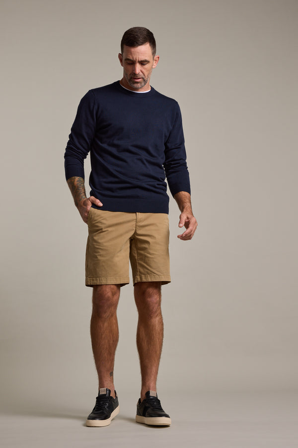A man stands against a plain background wearing a navy sweater, black sneakers, and Barkers Kingly Chino Short men's stretch shorts in tan, looking down with one hand in his pocket and a neutral expression.