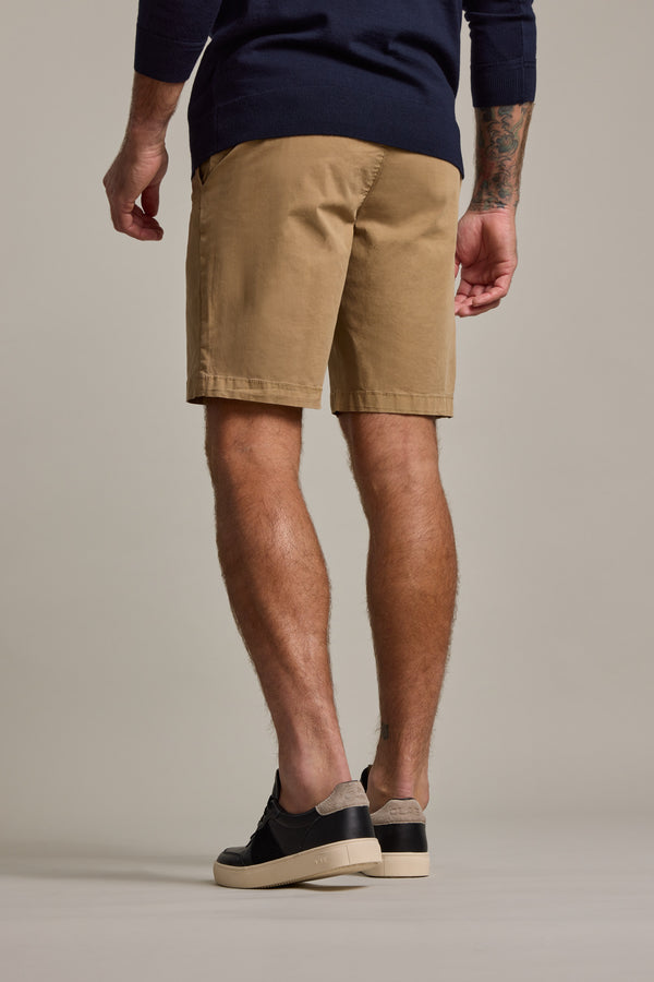 A person is seen from behind wearing Barkers Kingly Chino Short in tan, a navy top, and black low-top shoes with light soles. One arm has tattoos. The backdrop is plain and neutral.