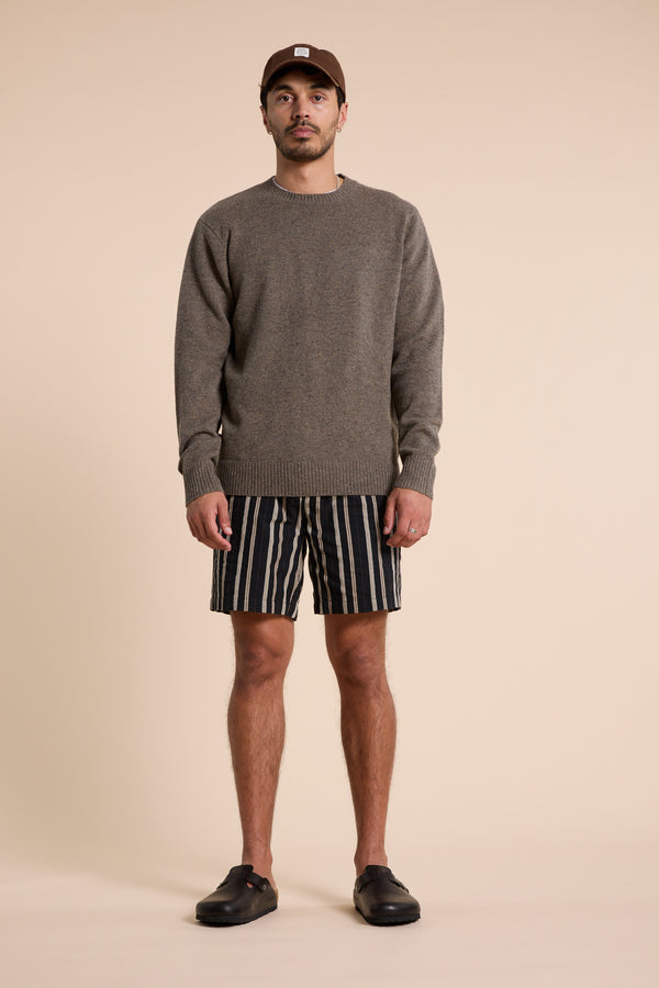 A man poses against a beige background, wearing Barkers Lawton Stripe Shorts with an elastic waistband, a brown sweater, black slip-on shoes, and a brown cap. He faces the camera directly with a neutral expression.