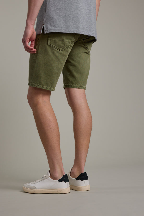 A person stands facing away in a neutral studio, wearing Barkers Rover Coloured Denim Short in olive green, a gray T-shirt, and white sneakers with black heel accents.