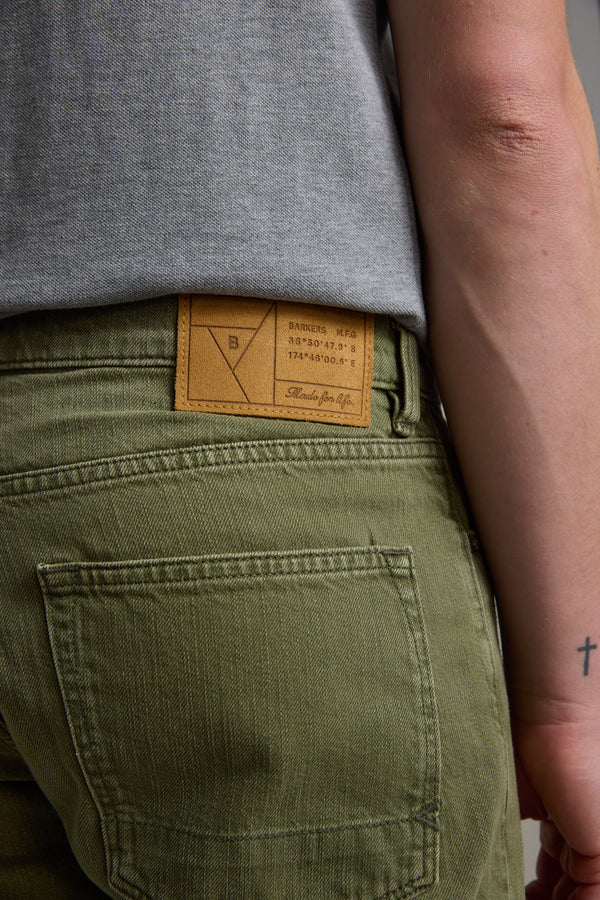 A person wears Barkers Rover Coloured Denim Shorts in olive green, featuring a brown leather waistband patch, paired with a gray shirt. Their forearm shows a small cross tattoo near the wrist.