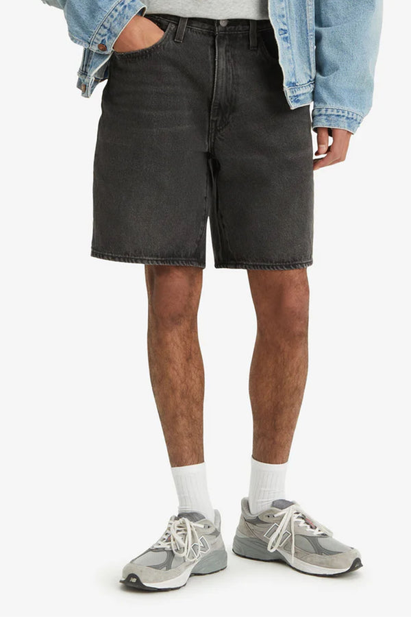 A person stands with one hand in their pocket, wearing Levi's 468 Loose Short by Levis in black non-stretch denim, a light gray t-shirt, light blue denim jacket, white socks, and gray sneakers. The image is cropped at the shoulders.