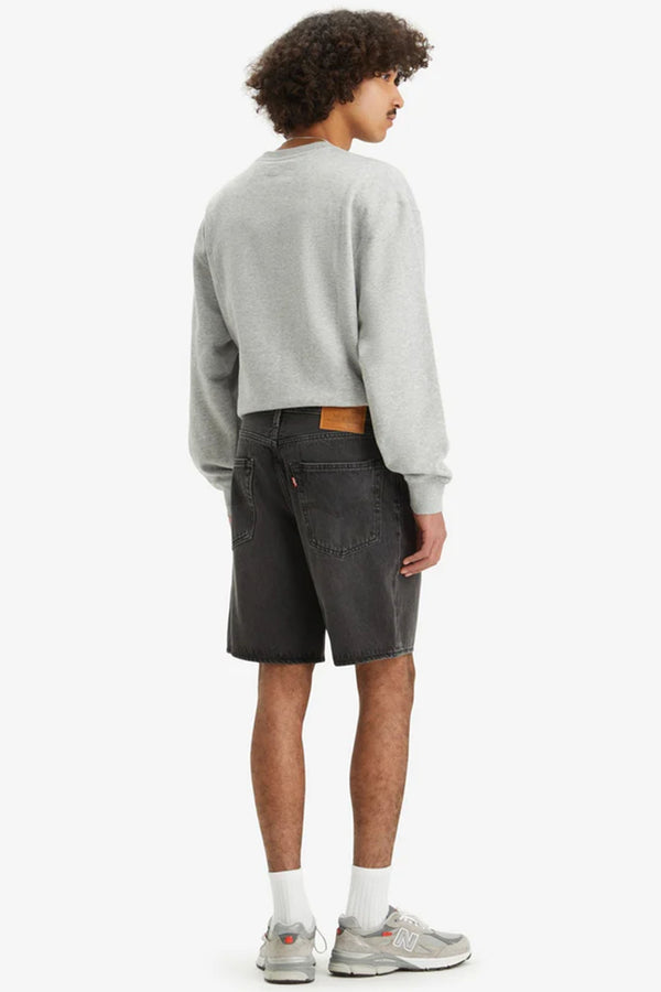 A person stands facing away, wearing a light gray sweatshirt, Levi’s 468 Loose Shorts in black non-stretch denim, white socks, and gray sneakers. The background is plain white.