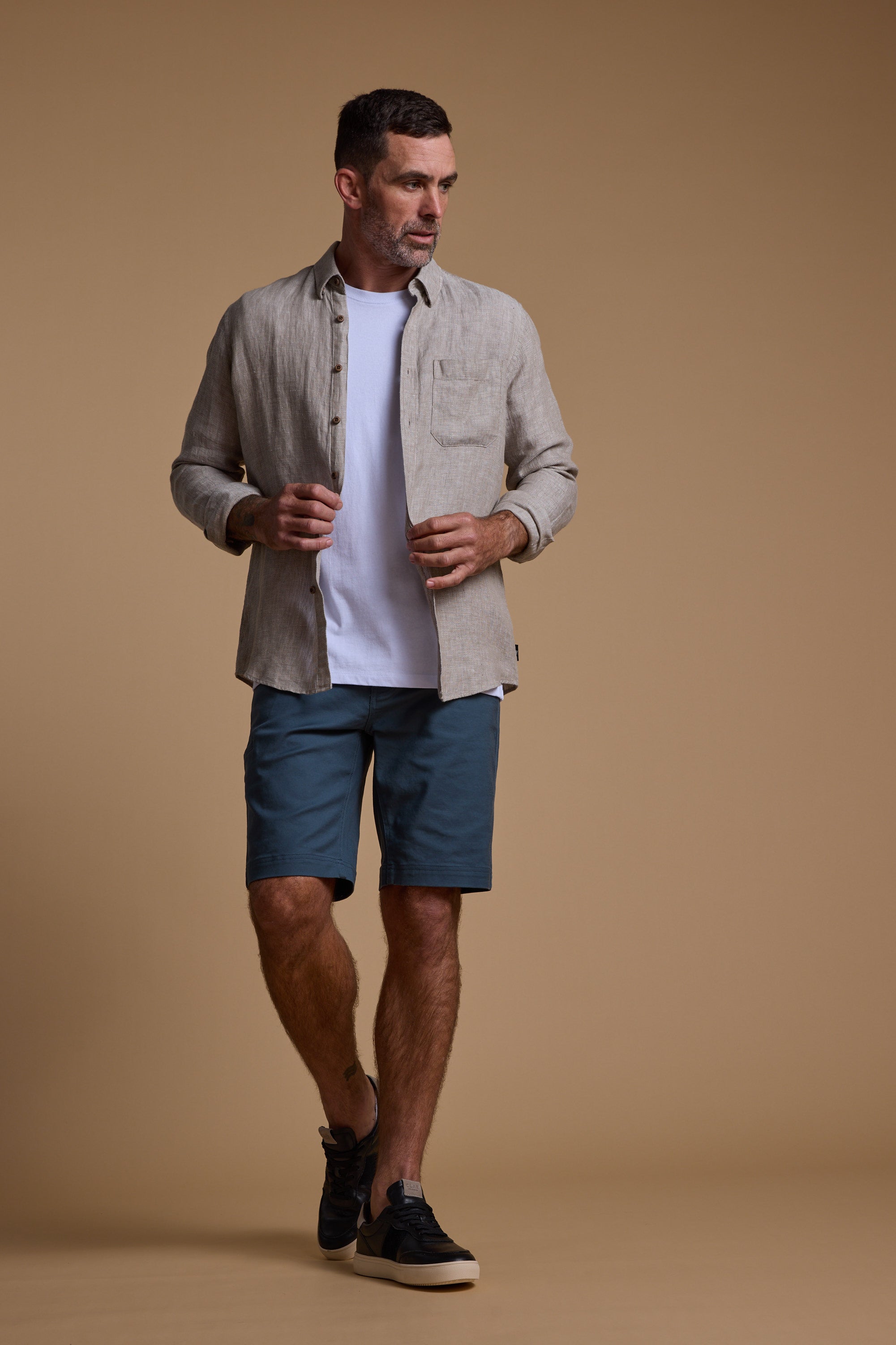 A man stands against a plain brown background wearing a tan button-up over a white tee, teal Barkers Anvil 5Pkt Shorts crafted from a durable cotton blend, and black sneakers. He looks to the side while adjusting his shirt.