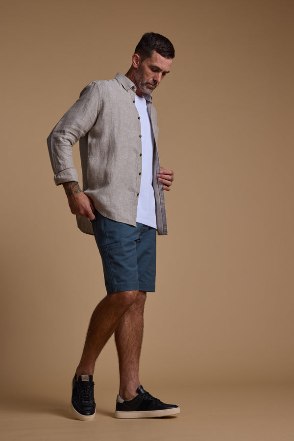 A man wearing a light beige button-up over a white tee, Barkers Anvil 5Pkt Shorts in teal, and black sneakers stands against a tan background, looking down with one hand in his pocket.