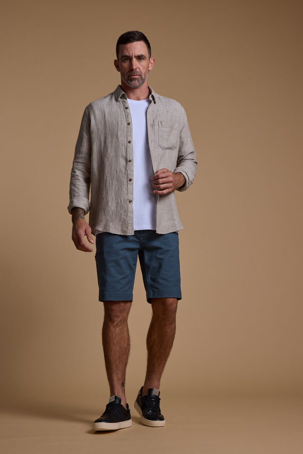 A man stands against a plain beige background wearing Barkers Anvil 5Pkt Short, styled with a light beige button-up over a white t-shirt and black sneakers. He looks forward with a neutral expression.