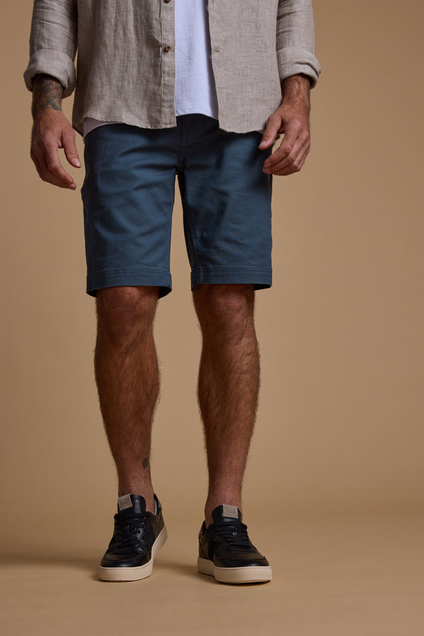 A man stands against a beige background wearing a light shirt, white T-shirt, Barkers Anvil 5Pkt Short in blue—a durable cotton blend—and black sneakers with white soles. His hands rest at his sides; only his body from the shoulders down is shown.