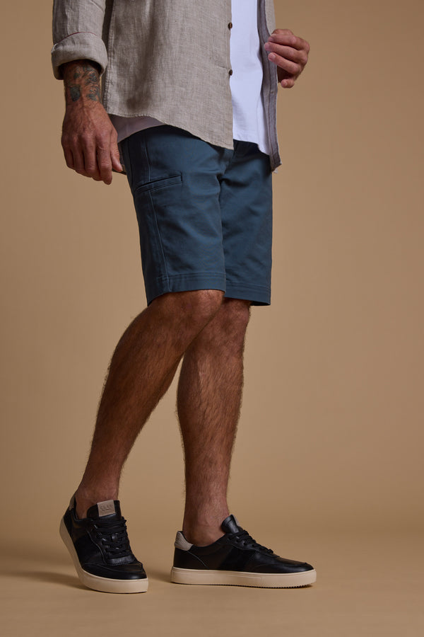 A man stands against a tan background wearing a Barkers Anvil 5Pkt Short in a durable cotton blend, beige shirt, white t-shirt, and black sneakers. His left arm shows visible tattoos. The photo is cropped at the shoulders.