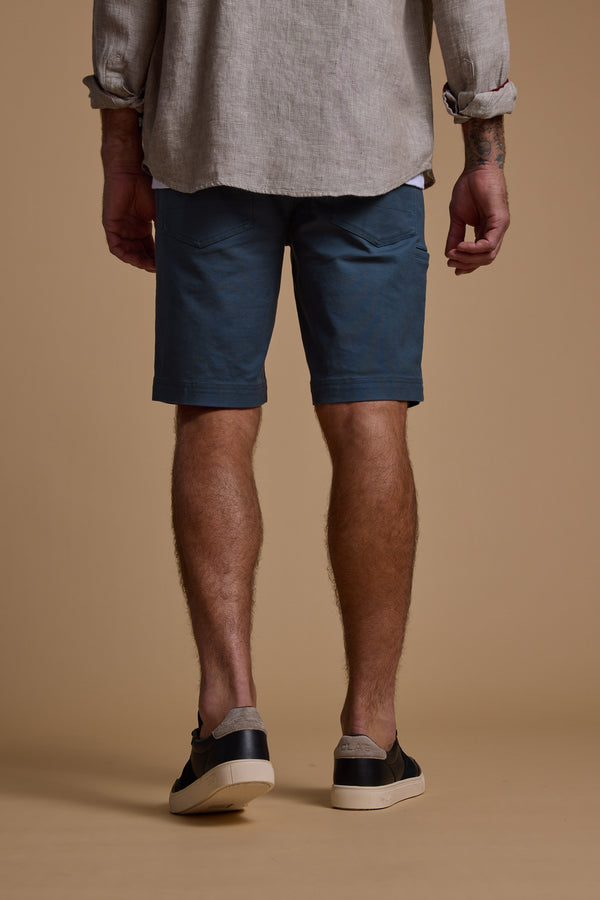 A man faces away, wearing a light gray long-sleeve cotton-blend shirt, Barkers Anvil 5Pkt Shorts in blue, and black slip-on shoes. His tattooed arms are revealed with rolled-up sleeves. The backdrop is plain beige.